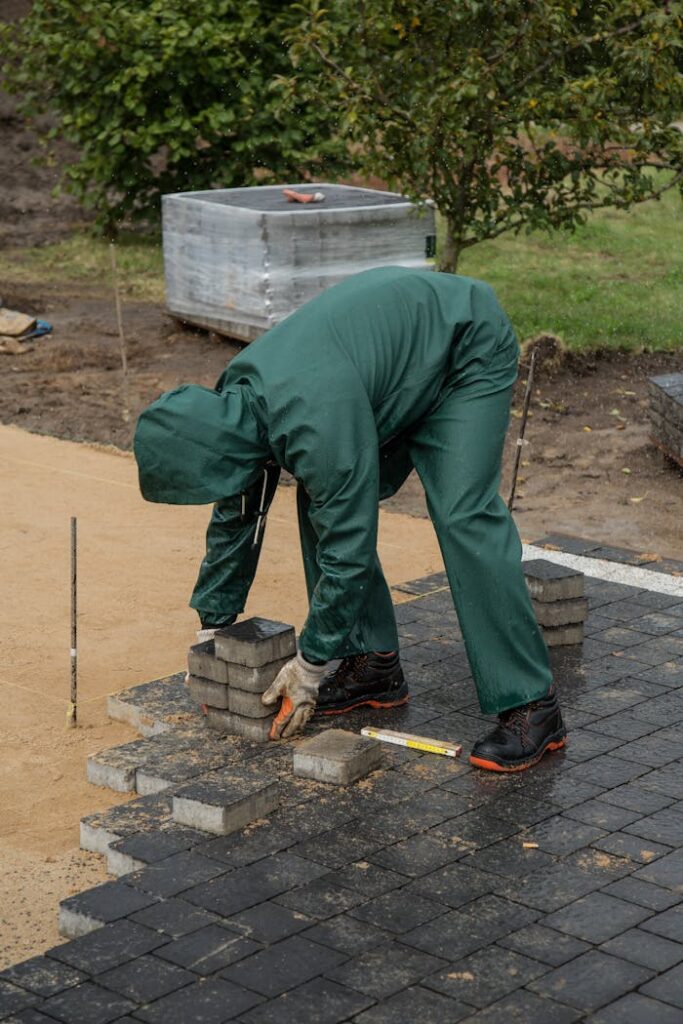 Man laying pavers in an outdoor setting, wearing green rain gear. Concept of construction work.